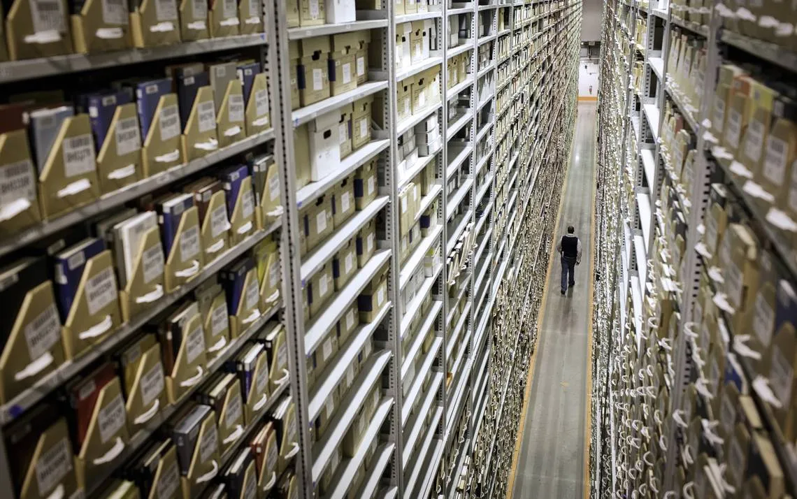Institutional archive — towering shelves of boxes stretching into the distance, a lone figure walking between them.